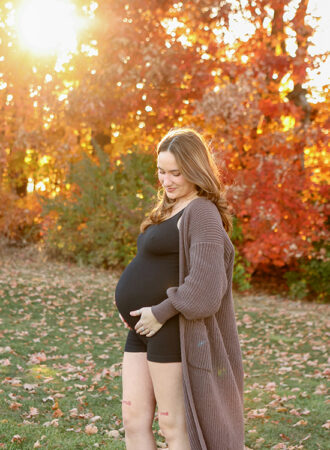 sun shines through Fall's golden trees behind woman cradling pregnant belly
