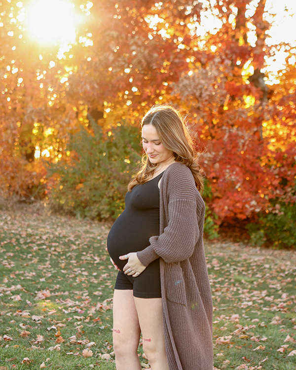 sun shines through Fall's golden trees behind woman cradling pregnant belly