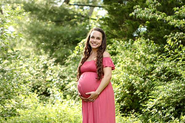 glowing pregnant woman looks down at her belly while standing in a shaded outdoor path